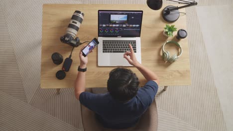 top view of a male color grading waving hand and having a video call on smartphone while sitting in the workspace using a laptop next to the camera editing the video at home