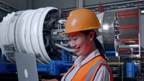 close up side view of asian female engineer with safety helmet working on a laptop while standing with airplane engine maintenance conducted