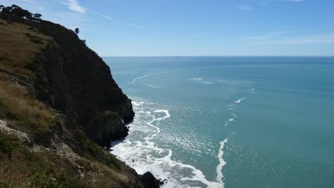 Cliff-face-shadow-contrasts-with-sunshine-on-beautiful-turquoise-ocean-as-waves-wash-onto-rocks---Godley-Head-Loop-Track,-Banks-Peninsula