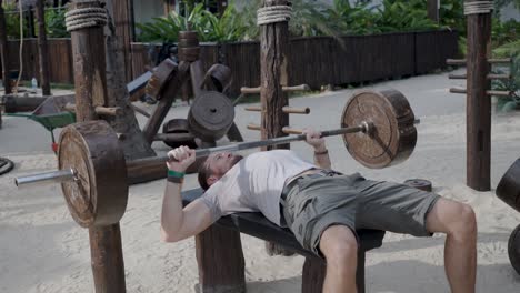 Caucasian male lifting weights, doing bench press, shoulder workout on the tropical thai beach in public outdoor gym.