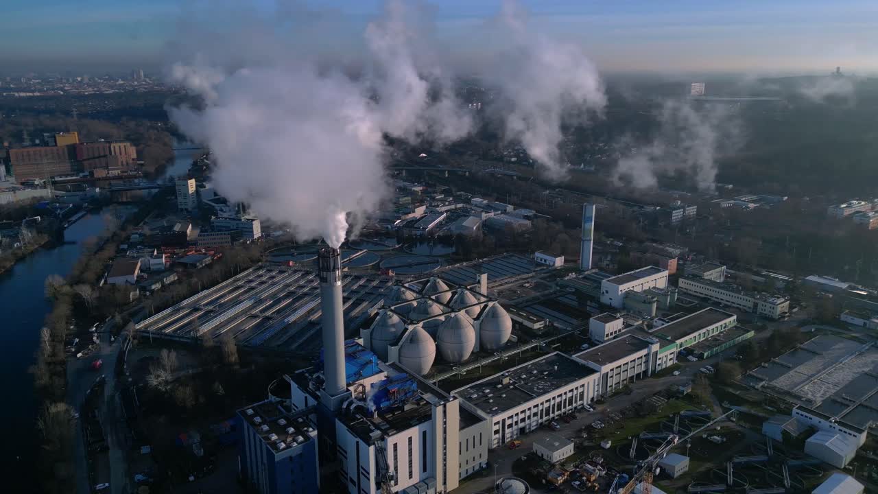 Industrial complex with a chimney emitting smoke into the sky, next to a sewage treatment facility and urban infrastructure. Perfect aerial view panorama orbit drone
