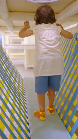 a small child walks carefully across a soft bridge trying to keep his balance, in an indoor soft playground with a distinctive blue and yellow striped net