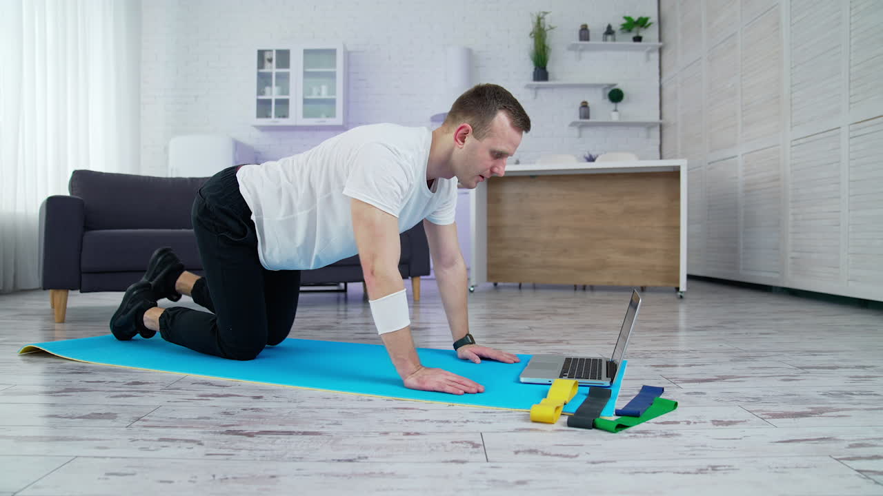 Guy in white t-shirt and black pants training in the kitchen. Young man doing exercise on a mat and looking into a laptop. Slow motion.