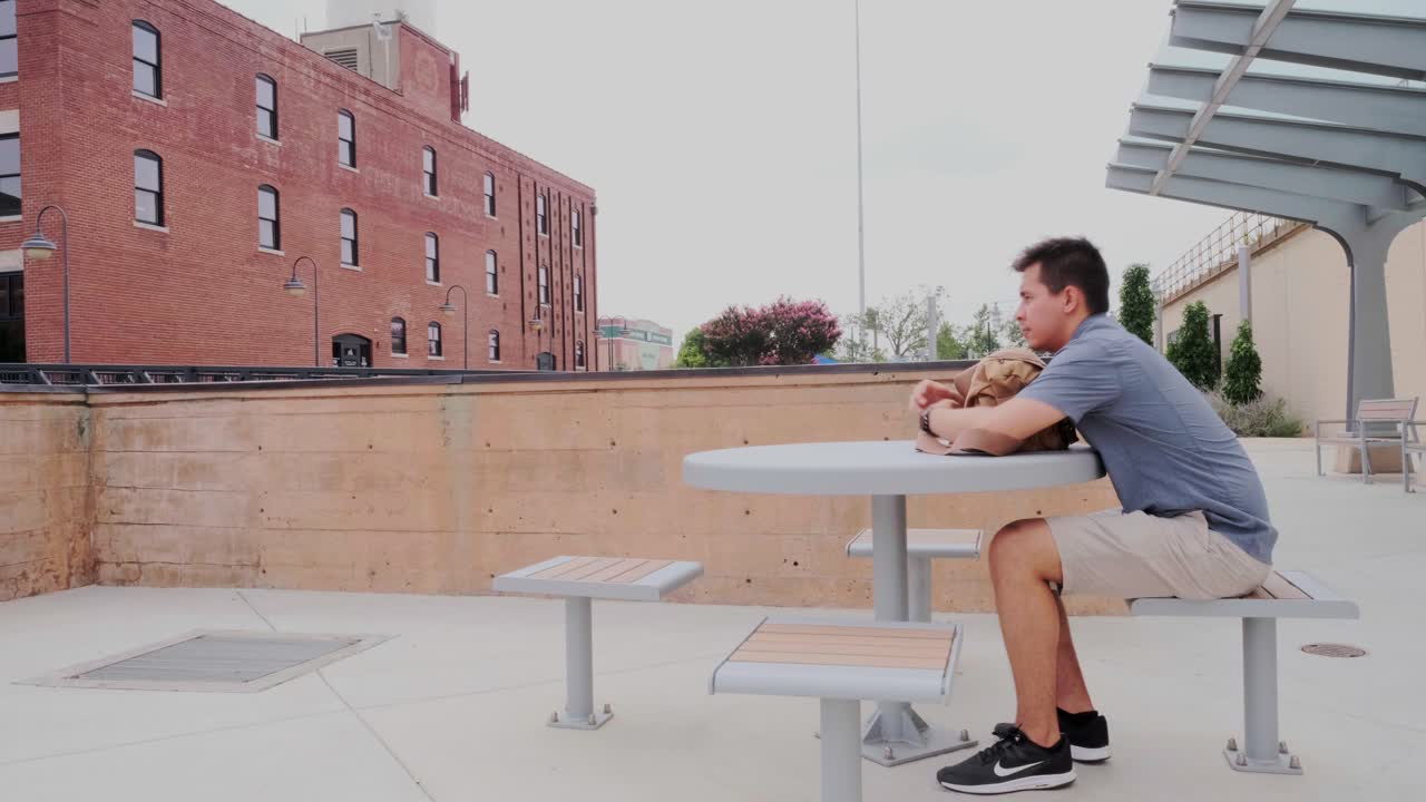 Wide view of young, male adult sitting and contemplating at public park table in Bricktown, Oklahoma City, USA. -static sho