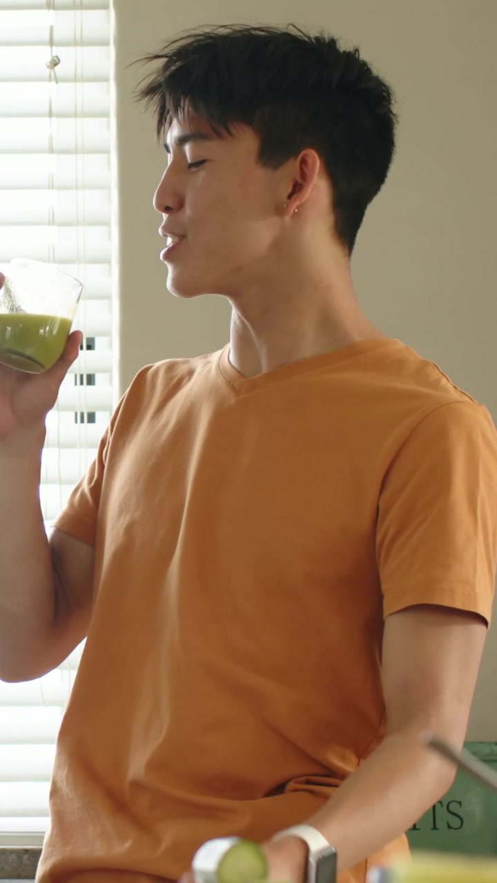 Video dọc: Người đàn ông mặc áo phông màu cam nhận ly được mời, nếm thử đồ uống màu xanh lá cây trong bếp tại nhà.
