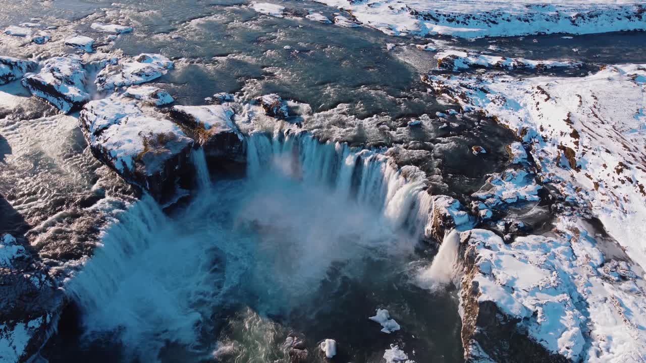 A dramatic aerial perspective showcases the powerful and iconic Godafoss waterfall, surrounded by snow and ice in the depths of winter