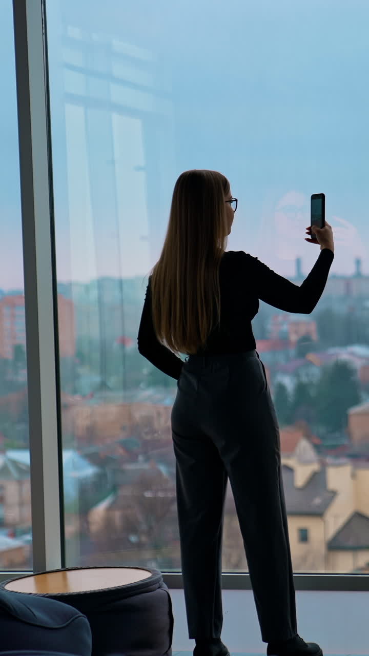 Long-haired girl with a phone indoors. Young woman shooting a video of a beautiful evening view of a city landscape from the office window. Rear view. Vertical video