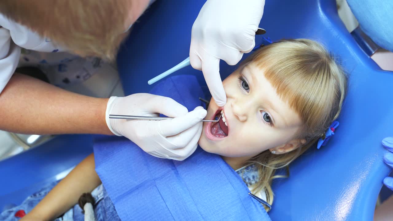 A dentist with a tool performs a medical examination of the teeth of a girl who is sitting in a blue chair