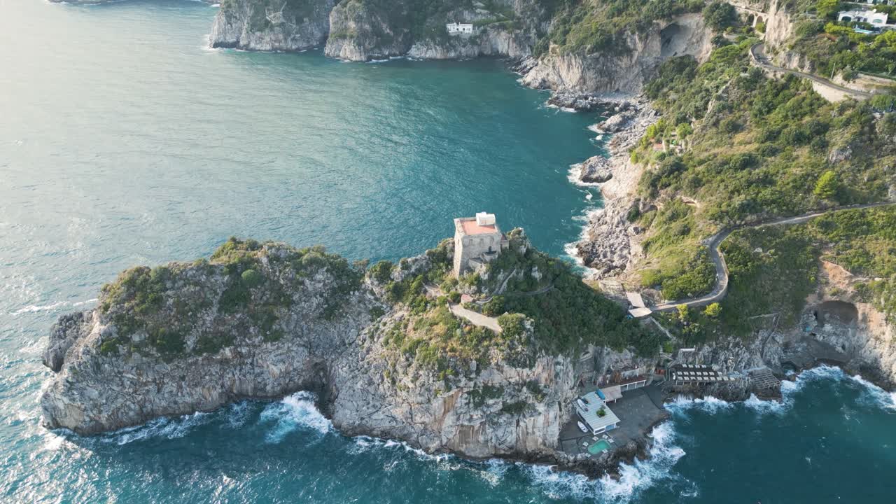 A breathtaking 4K aerial drone shot of the Amalfi Coast, Italy, featuring a secluded rocky peninsula with a historic coastal villa, turquoise waters, and dramatic cliffs.