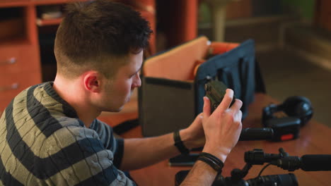 person sitting at table reaching into camera equipment bag. focus on hand, with camera, microphone, and smartphone visible on table. indoor relaxed environment with soft lighting and couch