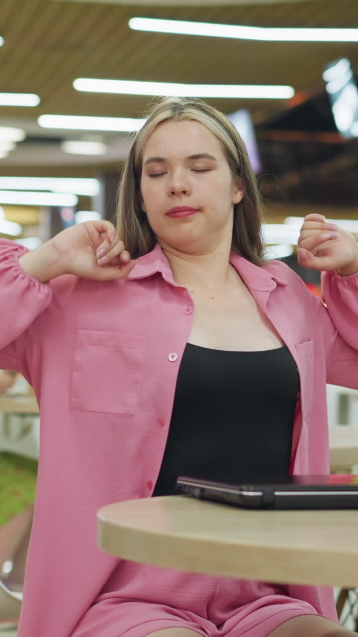 Lady in pink closes her laptop, looking bored and tired in a busy restaurant as she takes a deep breath and stretches, other people are seated in the restaurant, with a slight blur in the background