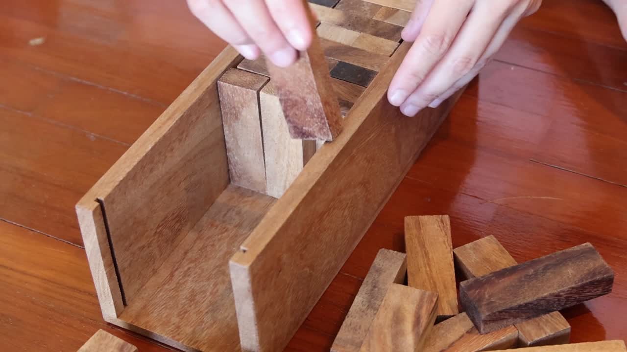 Hands skillfully place wooden blocks inside a rectangular wooden box on a table.