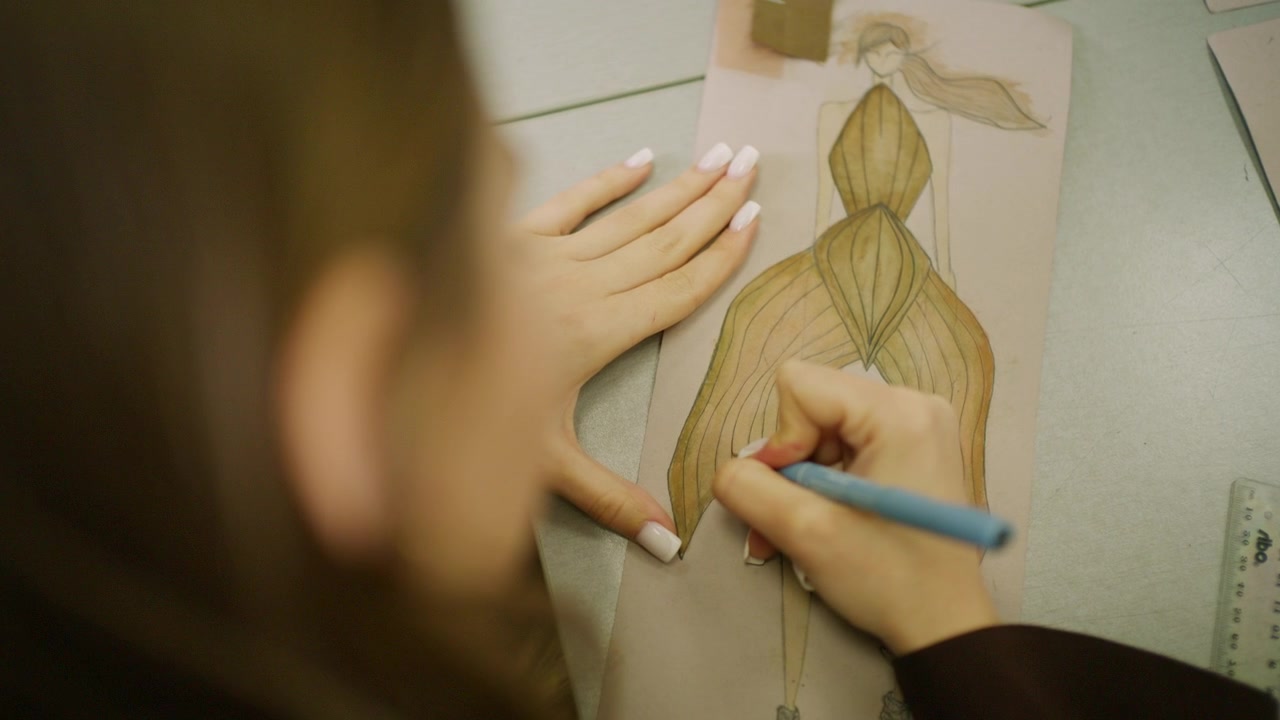 Primo piano di una stilista che disegna l'illustrazione di un abito strutturato dorato con matite colorate su un tavolo da lavoro in studio.