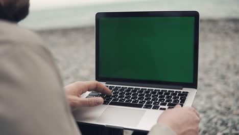 alone man is typing on a laptop with green-screen, sitting on seashore