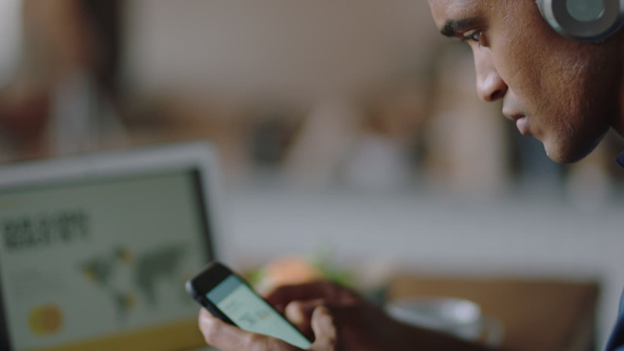 close up young businessman using smartphone in cafe drinking coffee browsing email messages networking online texting listening to music wearing headphones