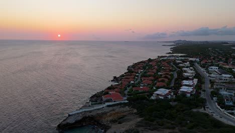 Serene-calm-tropical-coastline-homes-with-sun-setting-below-horizon-in-distance