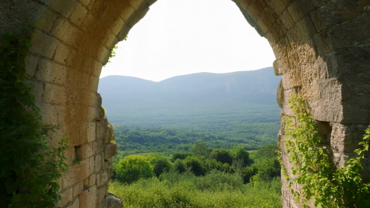 Camera advances toward weathered stone arch opening as bright daylight reveals climbing vines and dense woodland below, while blue-green mountains fade softly into atmospheric haze.