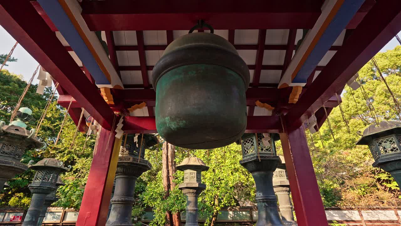A weathered bronze bell hangs beneath a red, traditional Japanese structure, surrounded by ancient stone lanterns. This close-up highlights the historical and spiritual essence of the shrine in Tokyo.