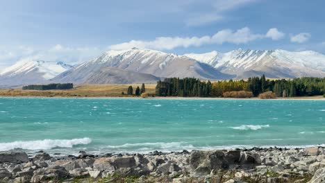 a wide, daylight panoramic shot slowly pans across a turquoise glacial lake, rocky shoreline, and distant snow-capped mountains under partly cloudy skies