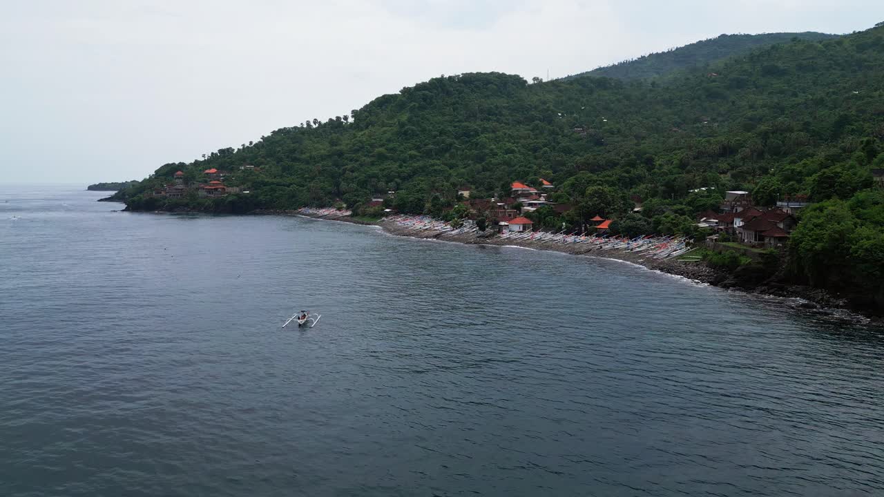 Imagens aéreas de uma vila de pescadores rural ao longo da costa, apresentando casas locais, colinas de floresta densa e um barco de pesca ancorado repousando na baía calma
