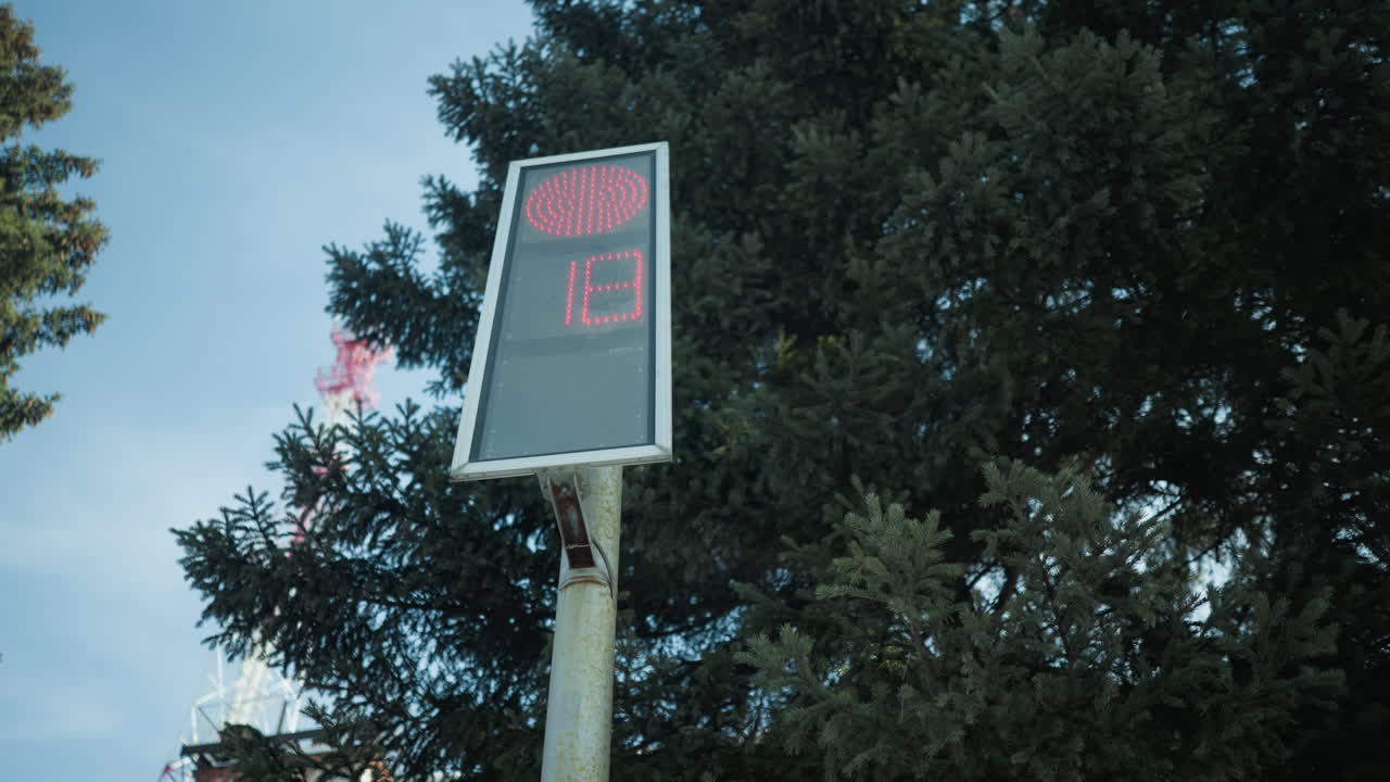 digital signpost with red led countdown reads 22 in winter light, metal pole before spruce canopy, telecom tower nearby, cold transit stop mood, quiet suburban scene under crisp sunshine