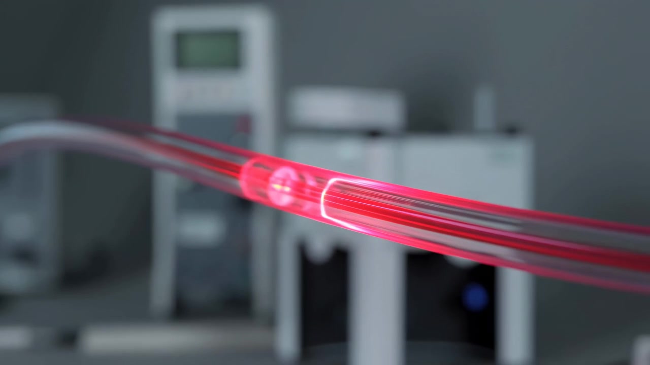 Vista orizzontale ravvicinata di tubi flessibili trasparenti che si inarcano su strumenti di laboratorio con una banda luminosa magenta concentrata lungo la curva centrale, riflessi morbidi, sfondo neutro e preciso