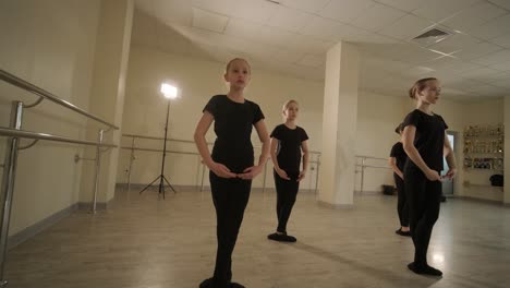 a group of young ballet students in black dancewear practicing positions in a spacious ballet studio with wooden flooring and wall-mounted barres. focused expressions and synchronized movements.