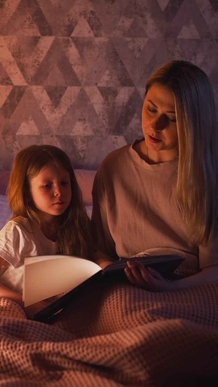 Careful long haired mother reads fairy tale to cute little daughter sitting together on bed with pink plaid in semidark children room slow motion