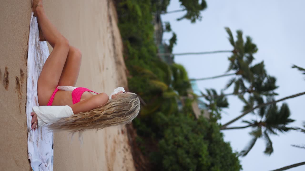 하와이 오아후 섬의 모래 해변에 앉아서 포즈를 취하는 분홍색 비키니를 입은 화려한 금발 여성의 세로 샷