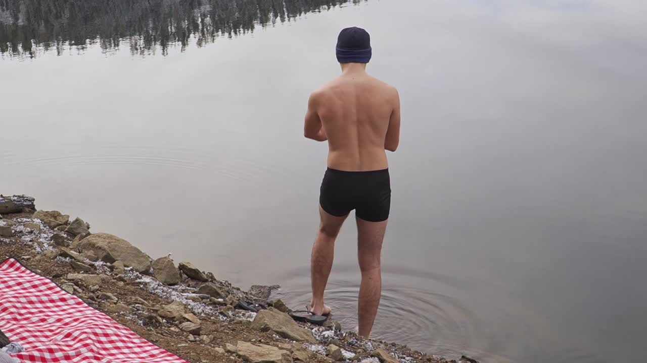 Man steps into still cold water of remote mountain lake while wearing only black swimming trunks and knit cap. Onsen, cold water therapy concept.