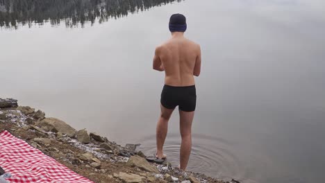 man steps into still cold water of remote mountain lake while wearing only black swimming trunks and knit cap. onsen, cold water therapy concept.