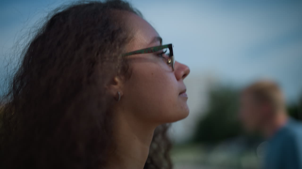 Close-up of lady with curly hair and glasses walking outdoors, adjusting hair backward while sunlight casts soft shadows on face, with blurred view of people and buildings in background
