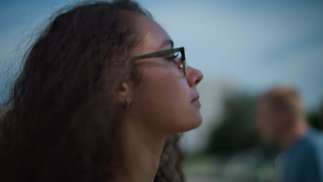 primer plano de una mujer con cabello rizado y gafas caminando al aire libre, ajustando el cabello hacia atrás mientras la luz del sol proyecta sombras suaves en la cara, con vista borrosa de personas y edificios en el fondo