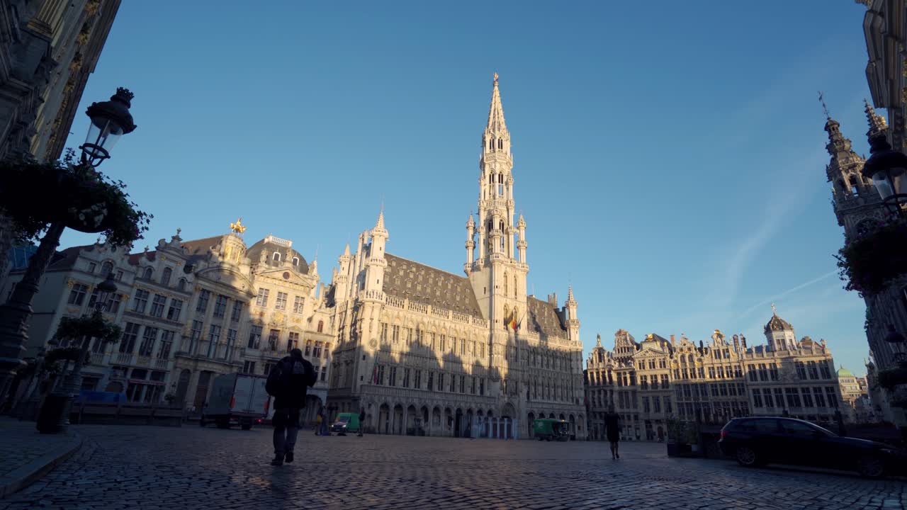 Grand Place Brussels, Belgium, wide static to shot sideways with town hall in the center and silhouette of person walking. On a calm warm summer morning with clear blue skies during sunrise.