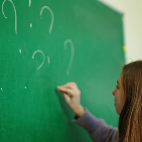 schoolgirl near the chalkboard. girl writing on green blackboard question marks in the classroom. education concept.