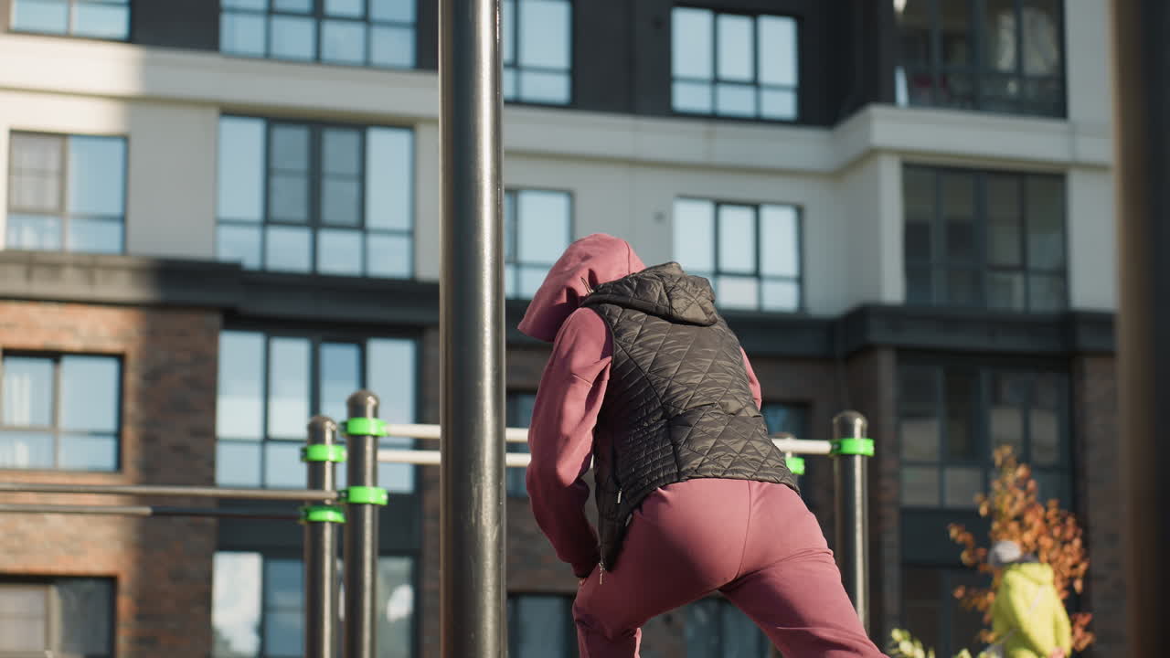 Back view youthful lady in sports wear doing split squat with hands braced on legs in urban park setting surrounded by soft morning sunlight, shadows on tile path, fitness and flowers blurred behind