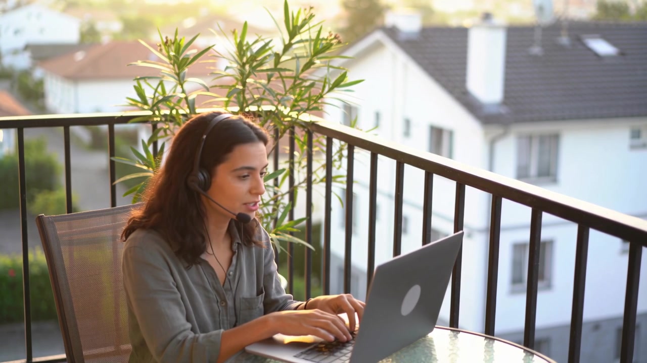 Typing woman in olive shirt turning to near sound, resuming headset call with laptop on balcony
