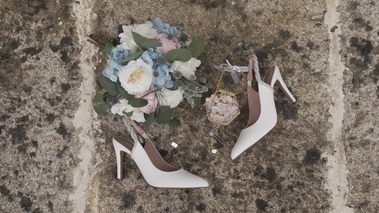 Eleganti accessori da sposa con tacchi bianchi, bouquet floreale e gioielli su pietra.