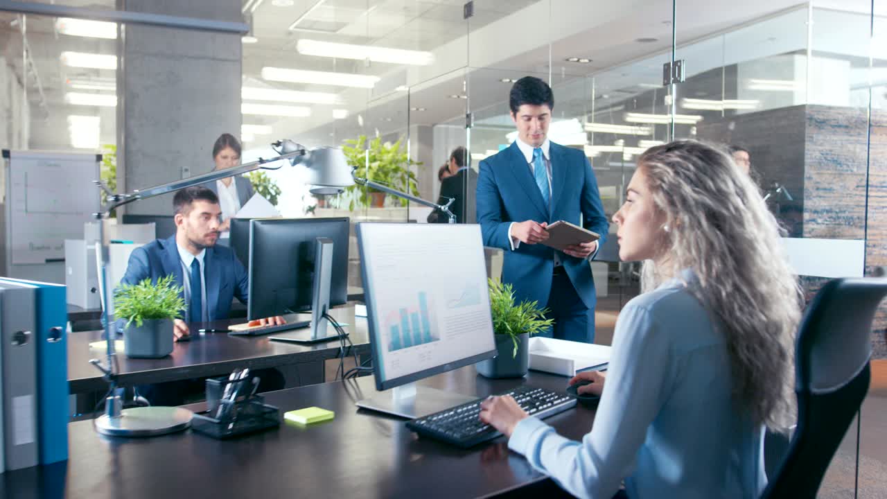 큰 사무실에서 동료는 그의 여성 임원에게 중요한 소식을 가져오고, 정보를 가진 태블릿 컴퓨터를 보여줍니다. 현대적인 유리와 콘크리트 건물은 바쁜 사업가들로 가득 차 있습니다.