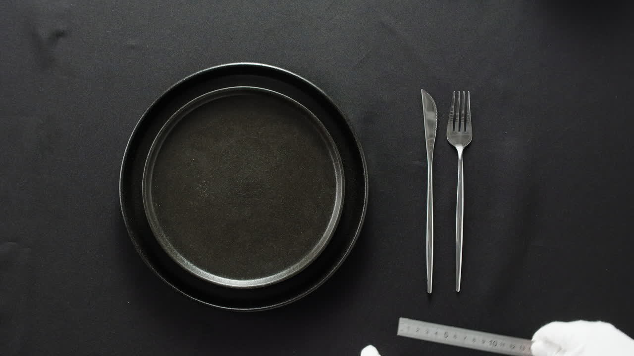 Minimalist table setting with white plate, fork, and knife arranged neatly, and a gloved hand adjusting cutlery.