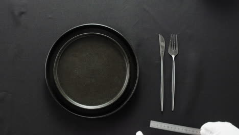 minimalist table setting with white plate, fork, and knife arranged neatly, and a gloved hand adjusting cutlery.