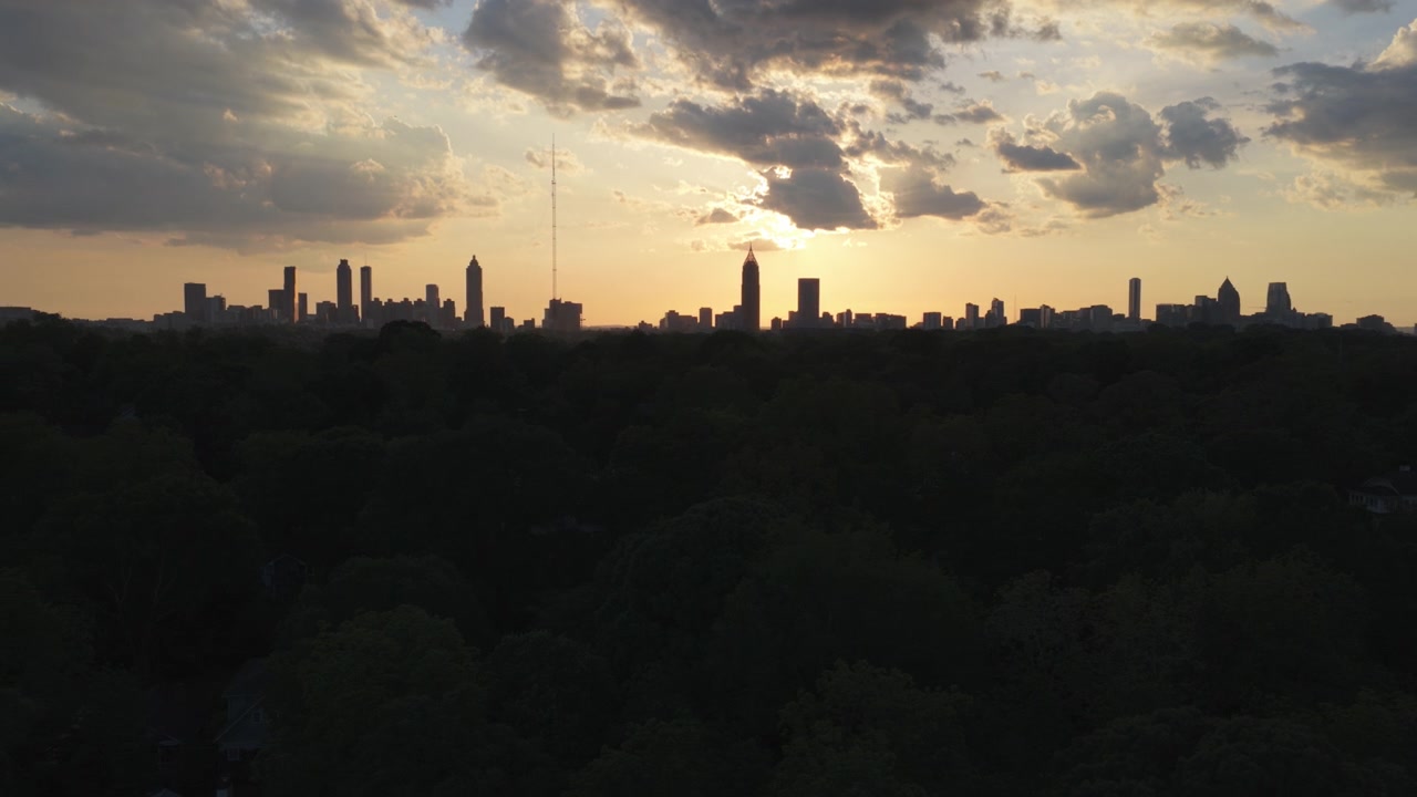 Una toma aérea cinematográfica al atardecer sobre un dosel arbóreo urbano de color verde oscuro hacia el icónico horizonte de Atlanta que se extiende por el horizonte durante una épica puesta de sol de verano con tonos naranjas y amarillos.