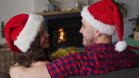 Rückansicht-Eines-Glücklichen-Kaukasischen-Paares-Mit-Weihnachtsmützen,-Das-Auf-Dem-Sofa-Sitzt
