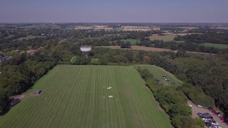 Vista-Aérea-De-Drones-Volando-Sobre-Un-Campo-Recién-Cortado-En-Trent-Park-Con-Estacionamiento-Y-Torre-De-Agua-A-La-Vista