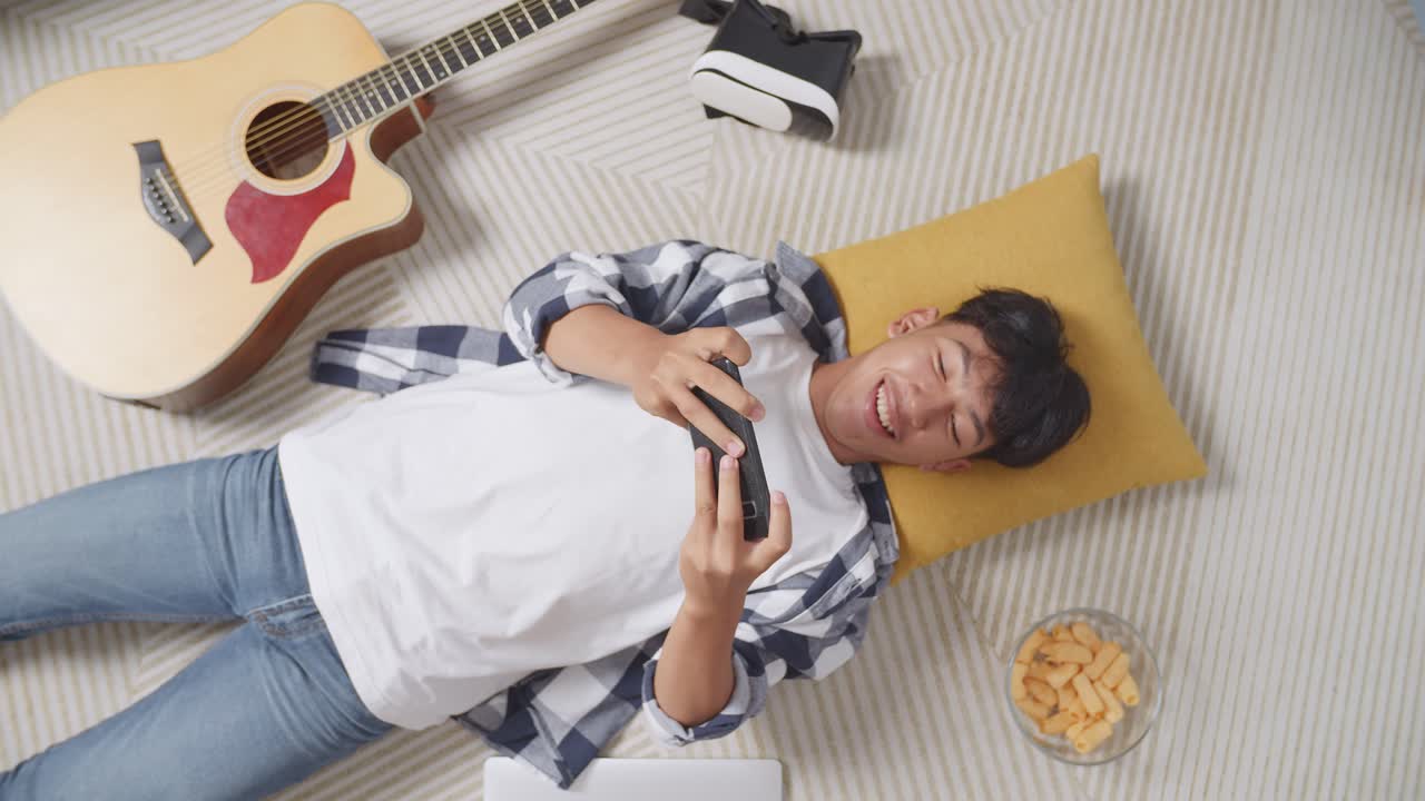 아시안 십대 소년이 집 바닥에 카에 누워 스마트폰으로 게임을 즐긴다.