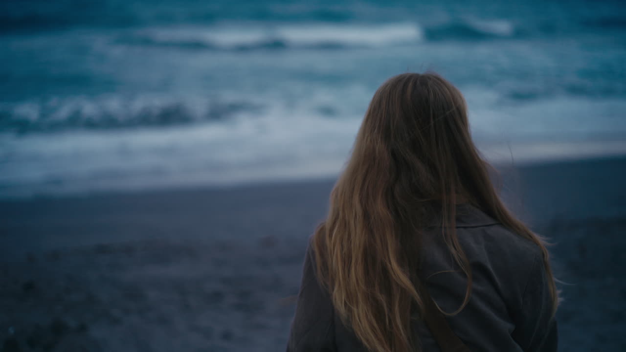 Vista posteriore di una giovane donna in piedi sulla spiaggia che guarda l'oceano durante l'ora blu, creando una scena costiera calma, riflessiva e suggestiva con luce soffusa e onde delicate
