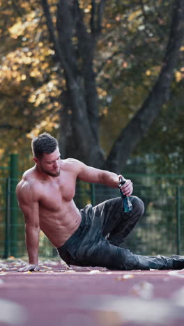 muscular sportsman outdoors. topless man with sportive body having break in the stadium. handsome athlete sits on autumn trees background. vertical video