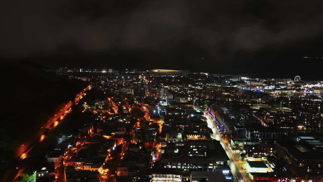 Cape Town shines at night as golden streetlights carve a glowing path along the hillside, overlooking the bustling city and harbor lights under a moody sky