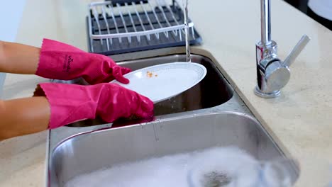 hands in pink gloves wash dishes under running water in a bright kitchen. the sequence captures the cleaning process with close-up shots