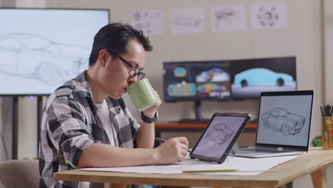 side view of asian male drinking coffee while drawing new car design concept on a tablet in the studio with tv and computers display 3d electric car model